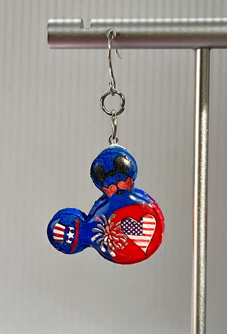 May include: A dangling earring featuring a blue, red, and white mouse-shaped charm. The charm displays a patriotic theme with a heart containing an American flag pattern, a firework burst, and a small top hat with stars and stripes on the ear.
