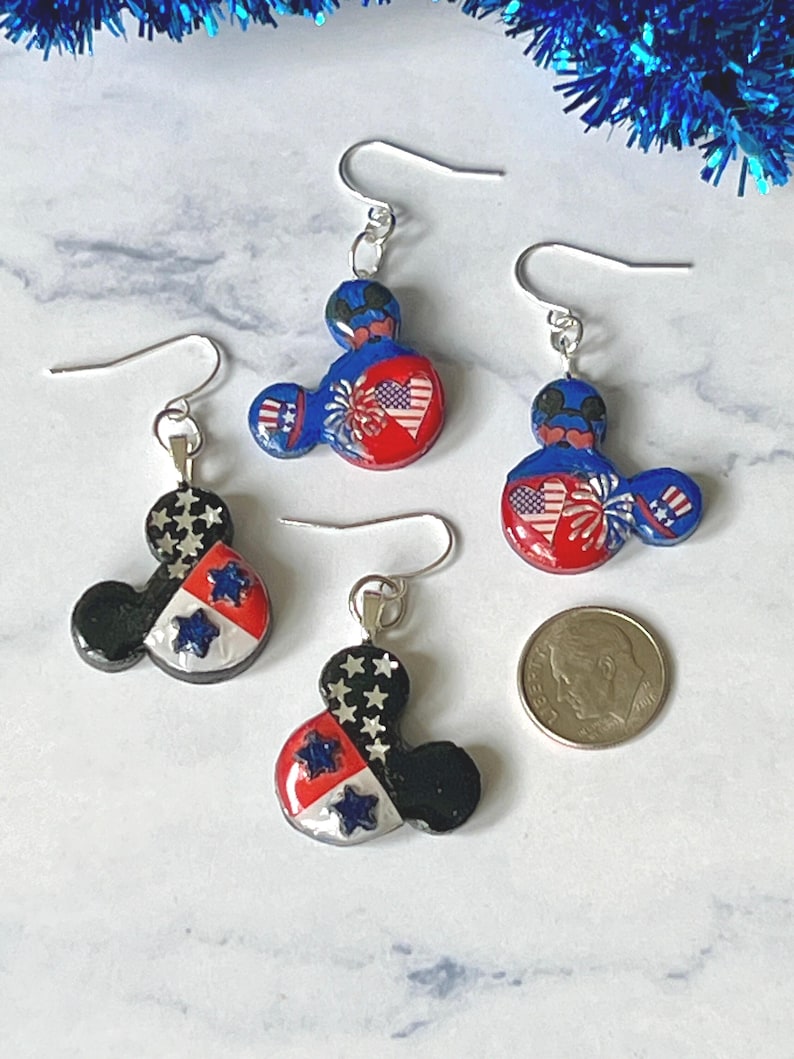 May include: Four dangle earrings shaped like mouse heads with silver-toned hooks. Two earrings feature red, white, and blue patriotic designs with hearts and fireworks. Two earrings feature black, white, and red designs with stars. A US dime is shown for scale.
