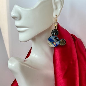 May include: Gold-tone dangle earrings with a whimsical design. The earrings feature three circular elements: a black bead, a button with a travel-themed image and the text "FAMILY VACATION", and a smaller gold-toned bead. The earrings are displayed on a white mannequin head.