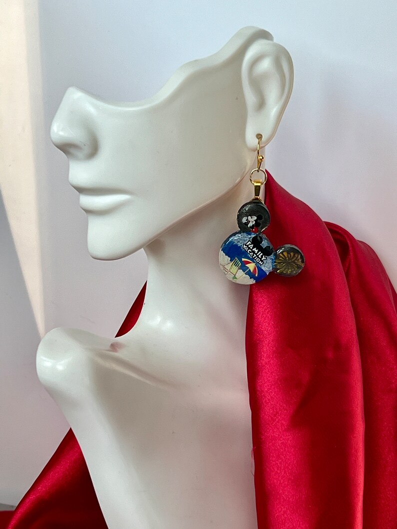 May include: Gold-tone dangle earrings with a whimsical design. The earrings feature three circular elements: a black bead, a button with a travel-themed image and the text "FAMILY VACATION", and a smaller gold-toned bead. The earrings are displayed on a white mannequin head.