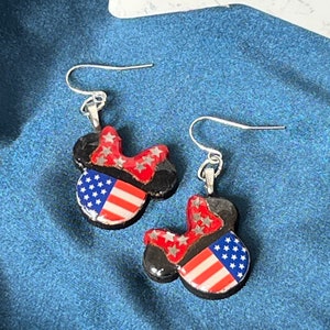 May include: A pair of dangle earrings featuring a black mouse-ear silhouette design. Each earring has a red bow with silver stars and a section patterned with red and white stripes and white stars on a blue background, attached to silver hooks.