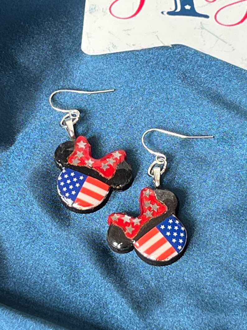 May include: A pair of dangle earrings featuring a black mouse-ear silhouette design. Each earring has a red bow with silver stars and a section patterned with red and white stripes and white stars on a blue background, attached to silver hooks.