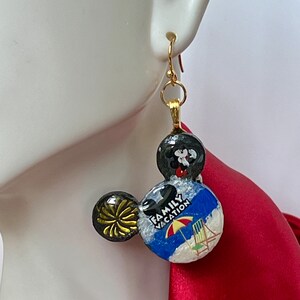 May include: Gold-tone Mickey Mouse-shaped earrings. One ear features a black and gold burst design, while the other displays a beach scene with the words "Family Vacation." The top sphere is black with a Mickey Mouse design.