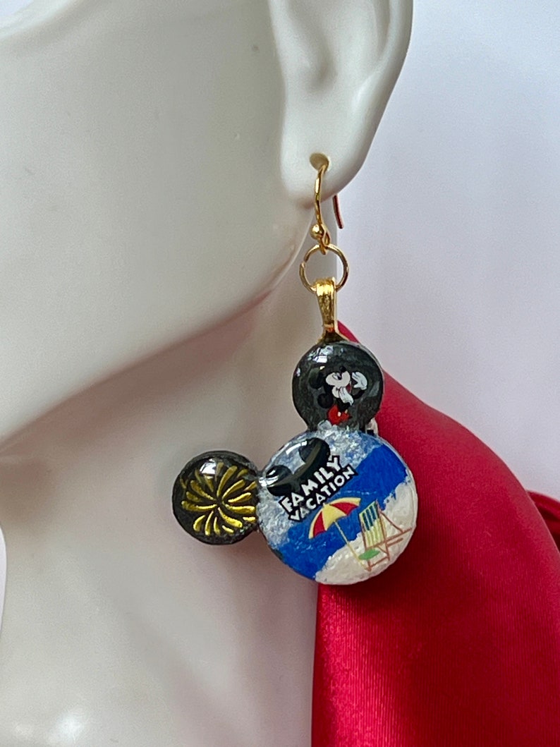 May include: Gold-tone Mickey Mouse-shaped earrings. One ear features a black and gold burst design, while the other displays a beach scene with the words "Family Vacation." The top sphere is black with a Mickey Mouse design.