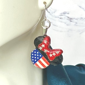 May include: A dangle earring featuring a black mouse-ear silhouette charm. One ear displays the American flag with white stars on a blue field and red and white stripes. The other ear is red with white stars, connected by a silver-tone metal hook.