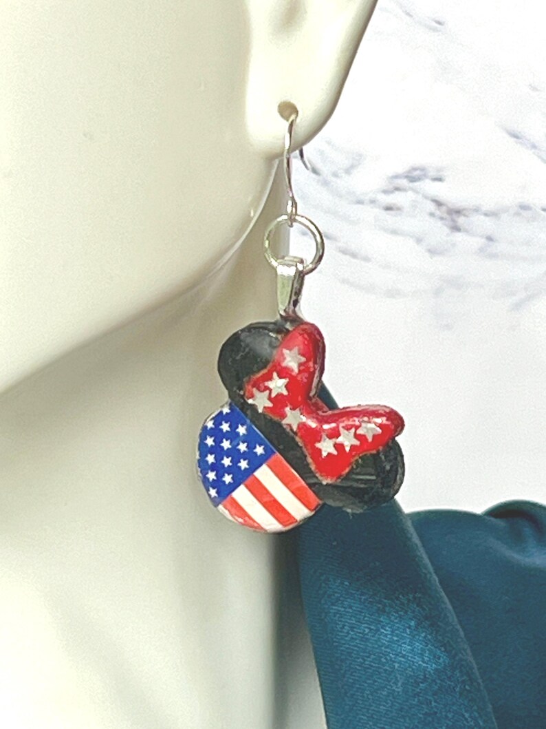 May include: A dangle earring featuring a black mouse-ear silhouette charm. One ear displays the American flag with white stars on a blue field and red and white stripes. The other ear is red with white stars, connected by a silver-tone metal hook.