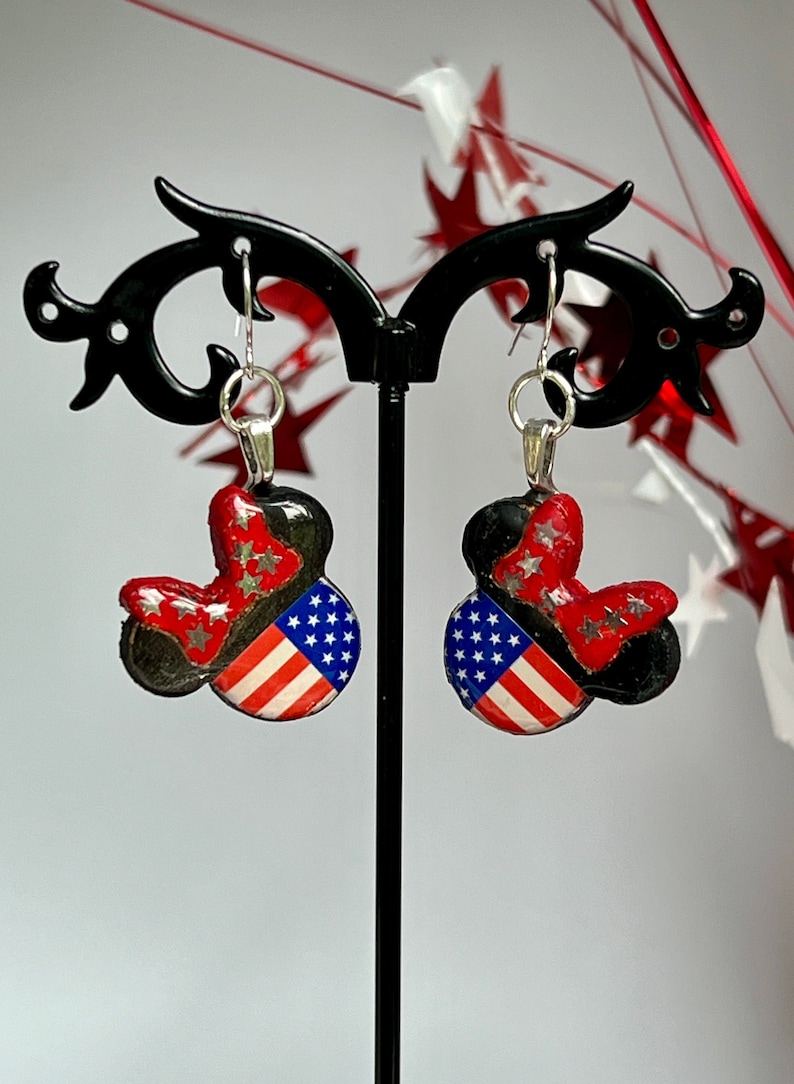 May include: A pair of dangle earrings featuring a mouse-shaped silhouette with a black base. The design includes a red bow with silver stars and a section patterned with the American flag, displaying white stars on a blue field and red stripes.