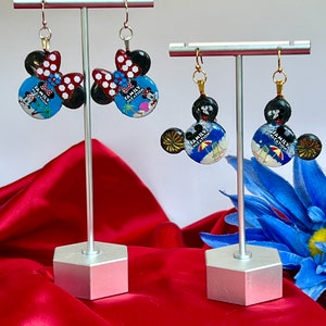 May include: Two pairs of Mickey Mouse-shaped earrings on display. One pair features a red and white polka dot bow, while the other has a black bow. The earrings have a blue background with the text "Family Vacation".