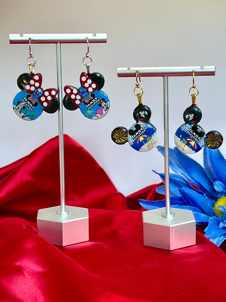 May include: Two pairs of Mickey Mouse-shaped earrings on display. One pair features a red and white polka dot bow, while the other has a black bow. The earrings have a blue background with the text "Family Vacation".