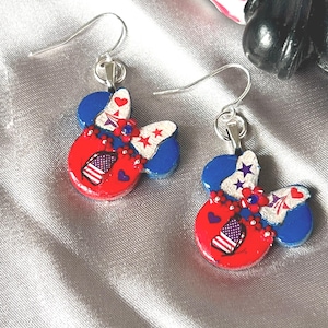 May include: A pair of dangle earrings featuring a mouse-ear silhouette in red, white, and blue. The design includes a bow with star and heart patterns, a small American flag motif, and floral accents, all suspended from silver-toned metal hooks.