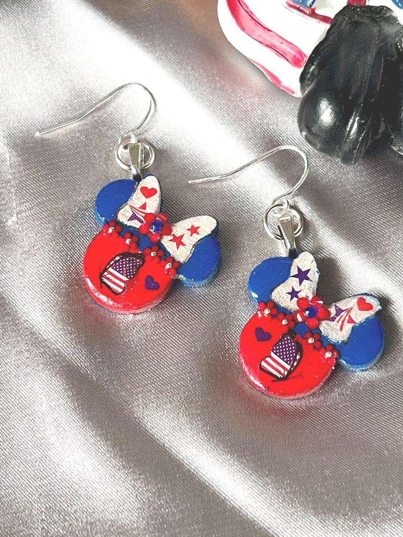 May include: A pair of dangle earrings featuring a mouse-ear silhouette in red, white, and blue. The design includes a bow with star and heart patterns, a small American flag motif, and floral accents, all suspended from silver-toned metal hooks.