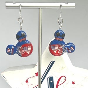 May include: A pair of dangle earrings featuring a mouse-shaped silhouette in red, white, and blue. The design includes a heart with an American flag pattern, firework graphics, and star motifs. The earrings hang from silver-toned metal hooks.