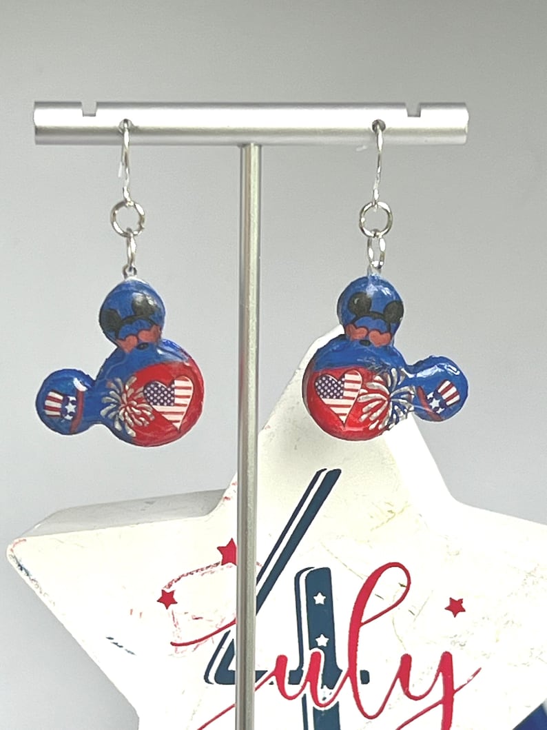 May include: A pair of dangle earrings featuring a mouse-shaped silhouette in red, white, and blue. The design includes a heart with an American flag pattern, firework graphics, and star motifs. The earrings hang from silver-toned metal hooks.