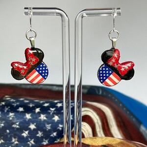 May include: A pair of dangle earrings featuring a mouse-ear silhouette. The design includes a black base, a red bow with silver star accents, and a section patterned with the American flag featuring white stars on a blue field and red and white stripes.