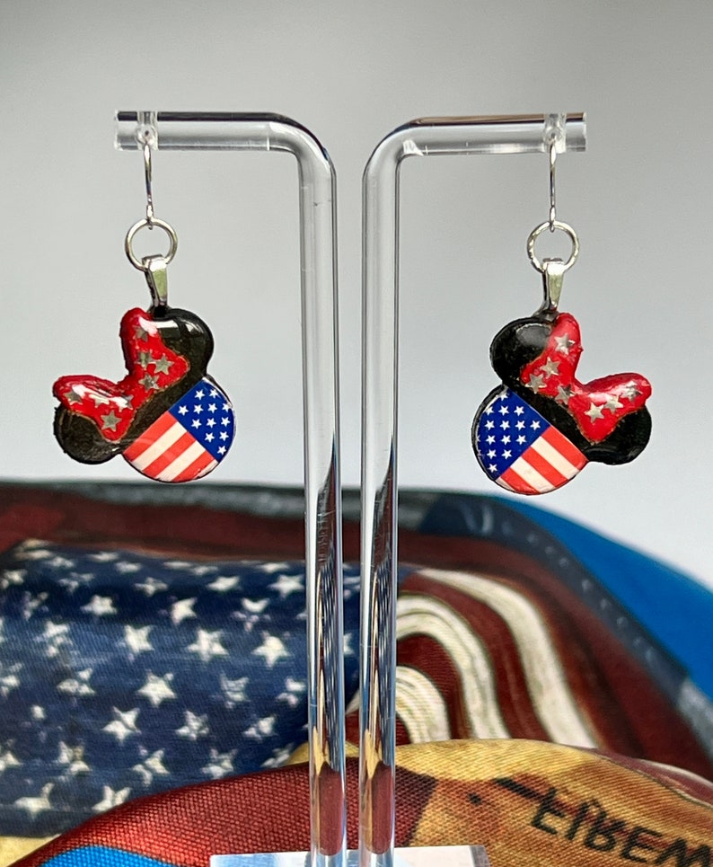 May include: A pair of dangle earrings featuring a mouse-ear silhouette. The design includes a black base, a red bow with silver star accents, and a section patterned with the American flag featuring white stars on a blue field and red and white stripes.