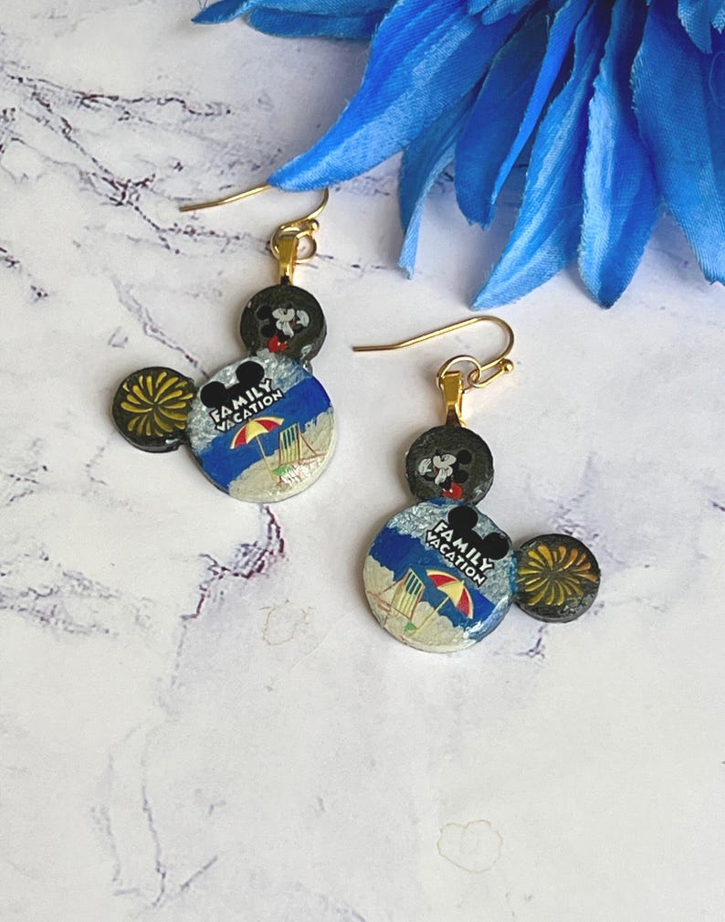 May include: Gold-tone dangle earrings featuring a Mickey Mouse design. The earrings have a black head and ears, with a circular beach scene that reads "Family Vacation". The earrings are a fun accessory.
