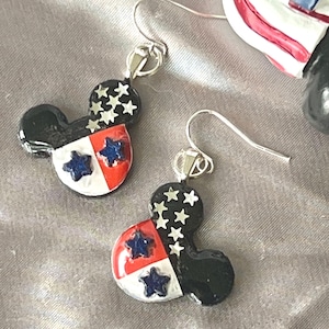 May include: A pair of dangle earrings shaped like mouse heads, featuring a black upper section with silver star accents and a lower section divided into red and white quadrants, each containing a dark blue star, attached to silver-toned metal hooks.
