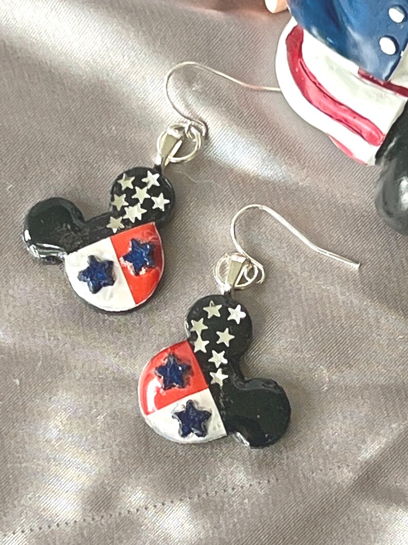 May include: A pair of dangle earrings shaped like mouse heads, featuring a black upper section with silver star accents and a lower section divided into red and white quadrants, each containing a dark blue star, attached to silver-toned metal hooks.