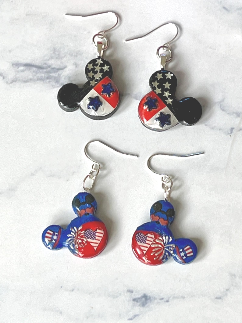 May include: Two pairs of dangle earrings featuring a mouse-ear silhouette. The top pair has a black, white, and red design with silver stars. The bottom pair features a blue and red design with heart-shaped American flags and firework motifs.