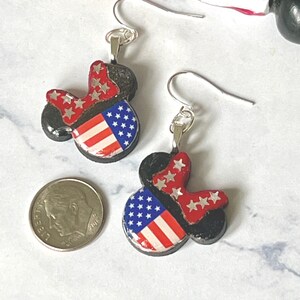 May include: A pair of dangle earrings shaped like mouse ears, featuring a red bow with white stars and a blue section with white stars and red and white stripes. A silver-colored US dime is placed next to the earrings for a size reference.