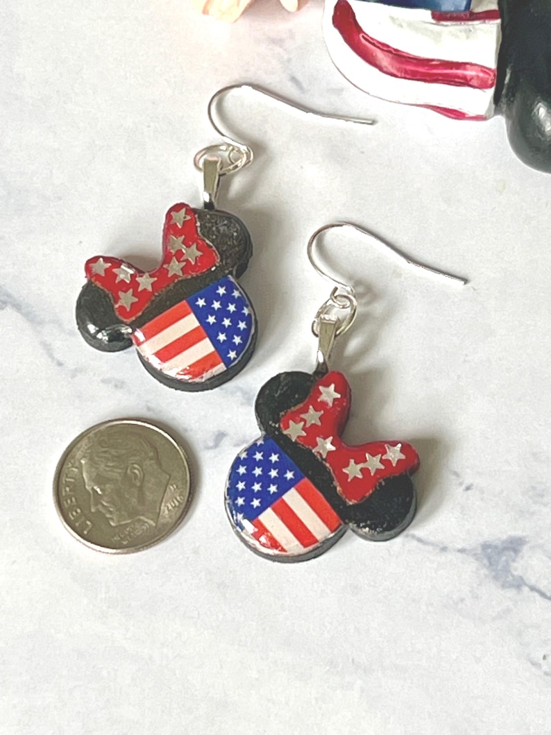 May include: A pair of dangle earrings shaped like mouse ears, featuring a red bow with white stars and a blue section with white stars and red and white stripes. A silver-colored US dime is placed next to the earrings for a size reference.
