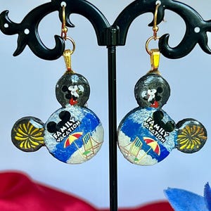 May include: A pair of Mickey Mouse-shaped earrings. The earrings feature a black Mickey Mouse head with a beach scene and the words "Family Vacation." The earrings have gold-colored hooks and accents.