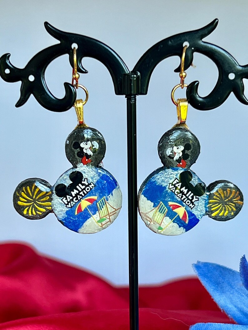 May include: A pair of Mickey Mouse-shaped earrings. The earrings feature a black Mickey Mouse head with a beach scene and the words "Family Vacation." The earrings have gold-colored hooks and accents.