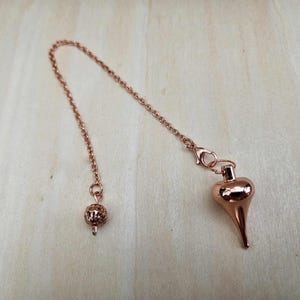 Pode incluir: Um p&ecirc;ndulo em ouro rosa com uma corrente e um peso em forma de gota. A corrente &eacute; presa a uma pequena bola decorativa em uma extremidade e a um peso em forma de gota na outra. O p&ecirc;ndulo &eacute; projetado para adivinha&ccedil;&atilde;o.