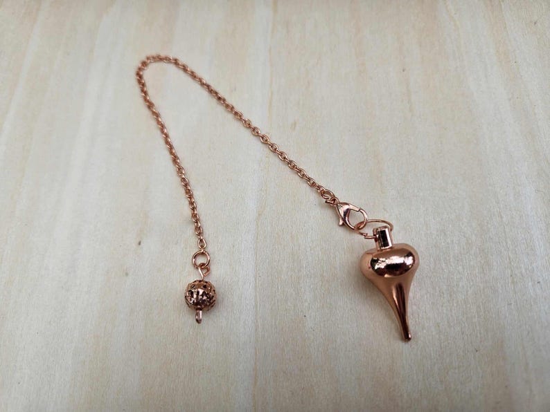 Pode incluir: Um p&ecirc;ndulo em ouro rosa com uma corrente e um peso em forma de gota. A corrente &eacute; presa a uma pequena bola decorativa em uma extremidade e a um peso em forma de gota na outra. O p&ecirc;ndulo &eacute; projetado para adivinha&ccedil;&atilde;o.