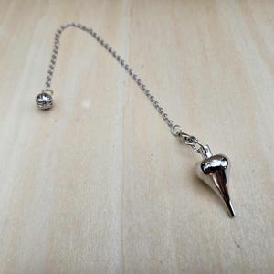 Pode incluir: Um p&ecirc;ndulo prateado com um peso em forma de gota pontiaguda, suspenso por uma corrente. A corrente conecta-se a uma pequena conta redonda no topo. O p&ecirc;ndulo est&aacute; sobre um fundo de gr&atilde;o de madeira clara.