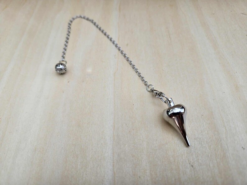 Pode incluir: Um p&ecirc;ndulo prateado com um peso em forma de gota pontiaguda, suspenso por uma corrente. A corrente conecta-se a uma pequena conta redonda no topo. O p&ecirc;ndulo est&aacute; sobre um fundo de gr&atilde;o de madeira clara.