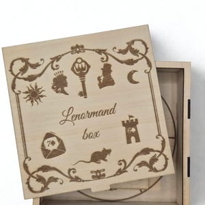 Peut inclure: Boîte en bois Lenormand avec des symboles gravés et une bordure décorative. Le couvercle ouvert présente une clé, un soleil et une lune. Le texte "Lenormand box" est gravé. Idéal pour ranger des cartes.