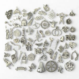 Puede incluir: Una colección de dijes plateados de varias formas, incluyendo una mariposa, una motocicleta, una brújula, un corazón, una balanza, un cohete y una pesa. Los dijes están dispuestos sobre una superficie blanca.