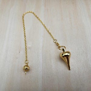 Pode incluir: Um p&ecirc;ndulo dourado com uma corrente e um peso em forma de gota. A corrente &eacute; presa a uma pequena conta redonda na outra extremidade. O p&ecirc;ndulo &eacute; projetado para adivinha&ccedil;&atilde;o e pr&aacute;ticas espirituais.