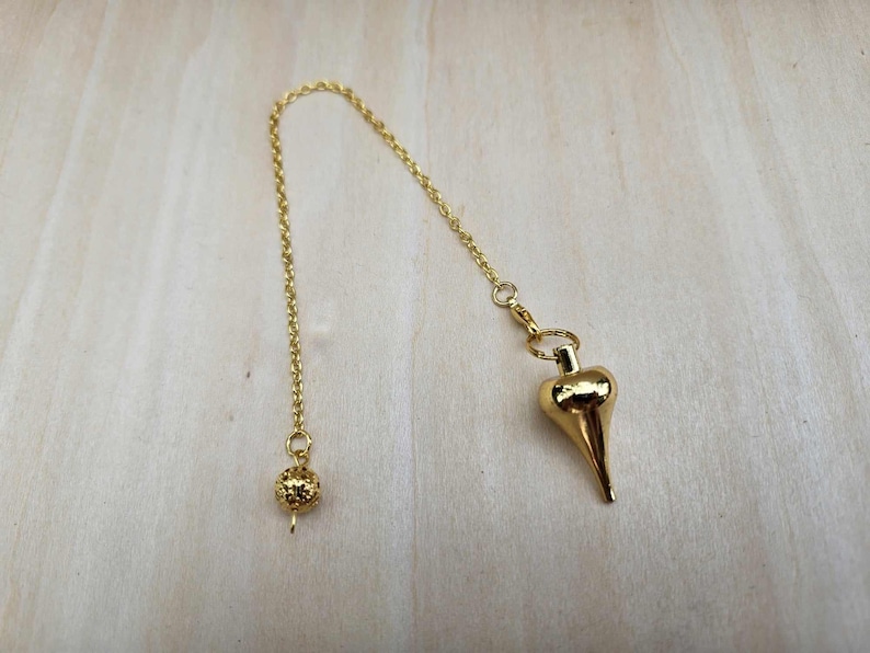 Pode incluir: Um p&ecirc;ndulo dourado com uma corrente e um peso em forma de gota. A corrente &eacute; presa a uma pequena conta redonda na outra extremidade. O p&ecirc;ndulo &eacute; projetado para adivinha&ccedil;&atilde;o e pr&aacute;ticas espirituais.