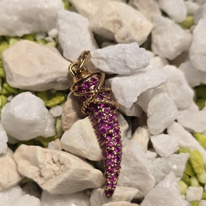 Könnte beinhalten: Ein goldfarbener Anhänger in Hornform, besetzt mit kleinen, leuchtend rosa Edelsteinen. Der Anhänger ist vor einem Hintergrund aus weißen und hellgrünen Kieselsteinen platziert, was einen auffälligen Kontrast erzeugt. Der hornförmige Anhänger ist ein Schmuckstück.