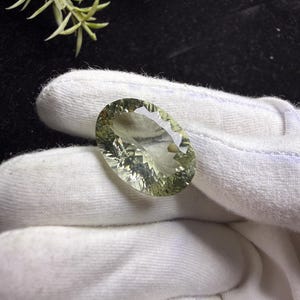 Könnte beinhalten: Ein oval geschliffener Edelstein mit einer blassgrünen, durchscheinenden Farbe. Der Stein weist einen Brillantschliff mit zahlreichen geometrischen Facetten auf, die das Licht reflektieren und eine funkelnde Optik auf der polierten Oberfläche erzeugen.