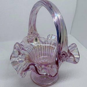 May include: A Fenton handmade iridescent glass basket in a soft lavender hue. The basket features a ruffled rim, a round base, and a twisted handle. The Fenton label is visible. The glass has a shimmering, opalescent quality.