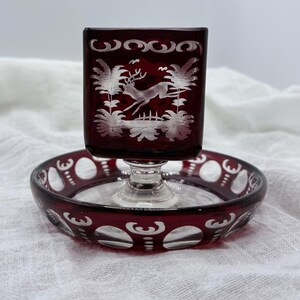 Puede incluir: Un plato y un soporte cuadrados de vidrio rojo rubí con un diseño grabado decorativo. El soporte presenta una escena de ciervo saltando. El plato tiene un diseño circular con patrones recortados. El vidrio es transparente.