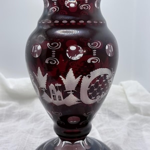 May include: A ruby red glass vase with intricate white etched designs. The vase features a flared top, a bulbous body, and a circular base. The etched patterns include swirls, dots, and a landscape scene. A decorative piece for home decor.