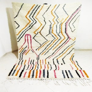 Puede incluir: Una alfombra marroquí blanca con un patrón geométrico en negro, rosa, naranja y gris. La alfombra tiene un borde de flecos.