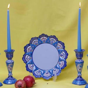 May include: A decorative arrangement featuring a round mirror with a blue and white floral border, flanked by two blue candlesticks with lit candles. Two pomegranates rest in front of the mirror. The background is yellow.