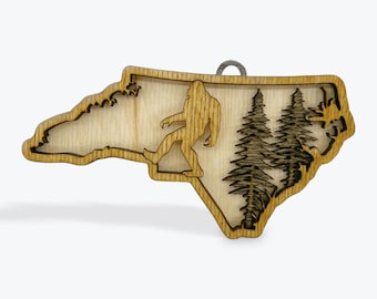 May include: A wooden ornament shaped like the state of North Carolina, with a silhouette of Bigfoot walking through a forest of pine trees.