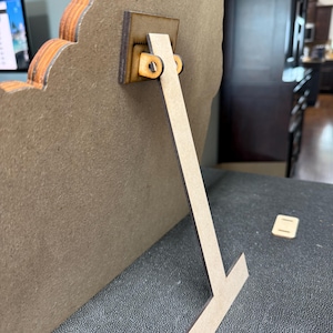 May include: A wooden picture frame with a decorative edge, supported by a wooden easel back. The easel back is attached with a small wooden bracket and two small wooden pegs. The frame is resting on a dark gray surface.