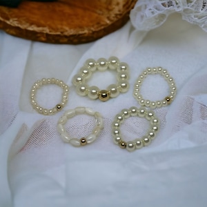 Puede incluir: Cinco anillos de perlas con detalles dorados. Los anillos están dispuestos sobre un fondo de tela blanca.