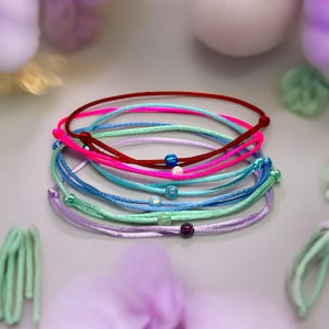 Puede incluir: Un conjunto de pulseras coloridas con pequeñas cuentas iridiscentes. Las pulseras están hechas de un cordón fino y sedoso y están disponibles en tonos de rojo, rosa, azul y verde.