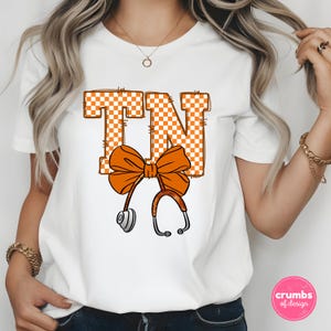 May include: White t-shirt featuring the letters "TN" in orange and white checkered pattern. An orange bow and stethoscope design is centered below the letters. The shirt is a short-sleeved crew neck.