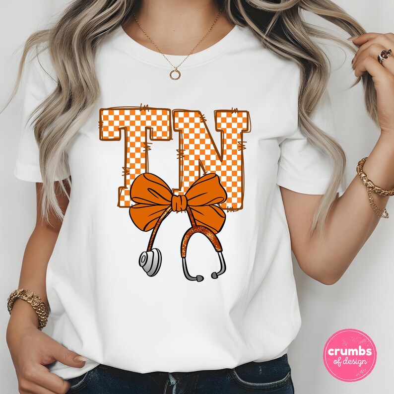 May include: White t-shirt featuring the letters "TN" in orange and white checkered pattern. An orange bow and stethoscope design is centered below the letters. The shirt is a short-sleeved crew neck.