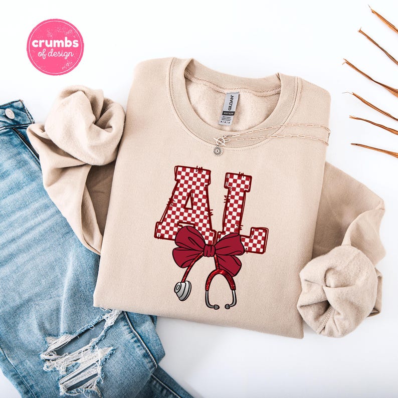 May include: A beige sweatshirt featuring the letters "AL" in a red and white checkered pattern, a red bow, and a stethoscope design. The sweatshirt is folded on a white surface with a pair of blue jeans.