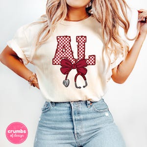 May include: Cream-colored t-shirt featuring the letters "AL" in a maroon and white checkered pattern, with a maroon bow and a stethoscope graphic. The shirt is paired with blue jeans.
